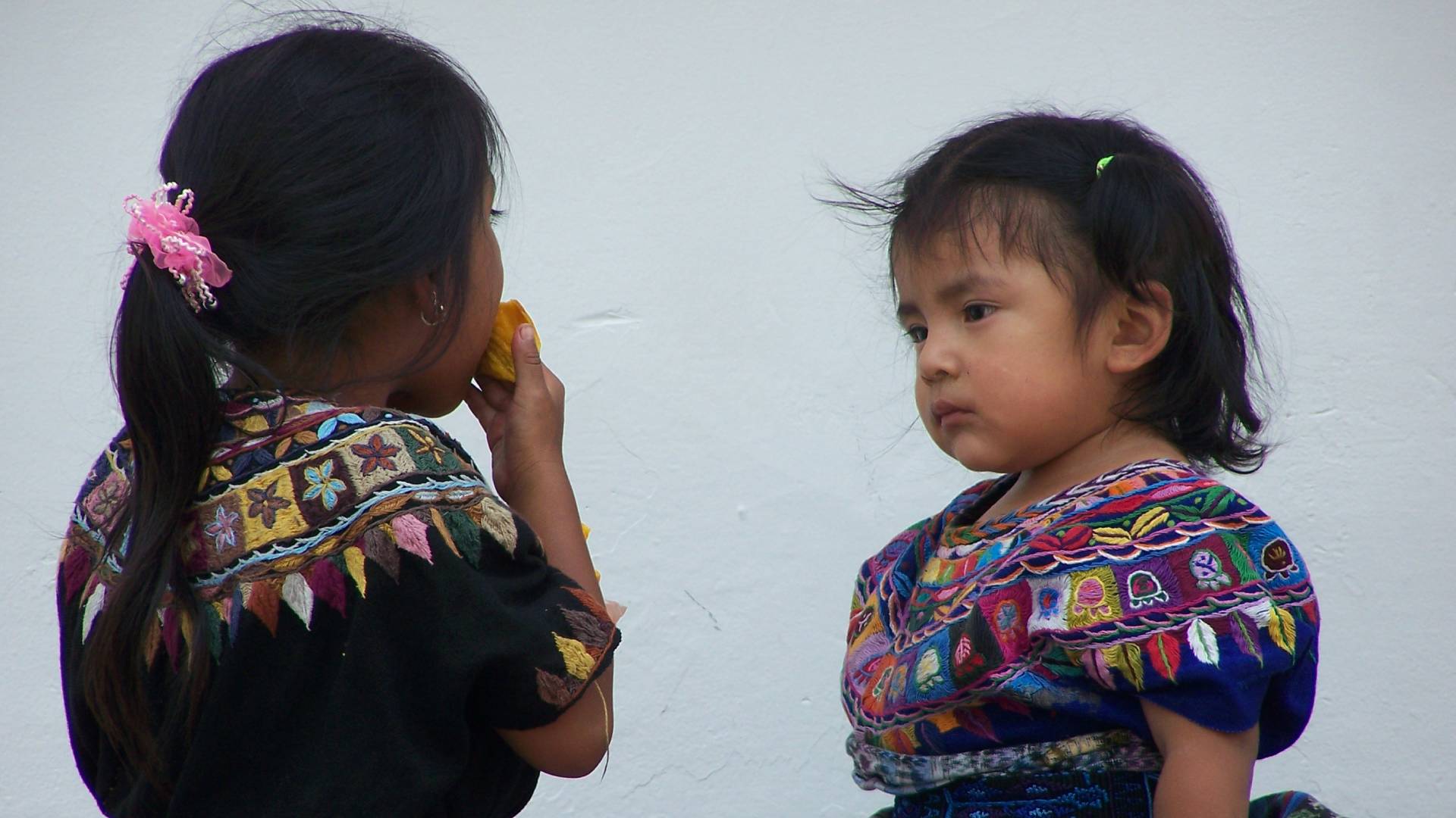 Two young girls are standing next to each other in this image. One girl is wearing a colorful dress and has a green hair clip, while the other girl has a pink flower in her hair. The girls appear to be talking or interacting with each other. Additionally, there is a person holding a banana in the background. The setting seems to be outdoors, possibly in a garden or park. The colors in the image are predominantly shades of pink, blue, and green. The girls seem to be enjoying themselves in each other''s company on a sunny day.