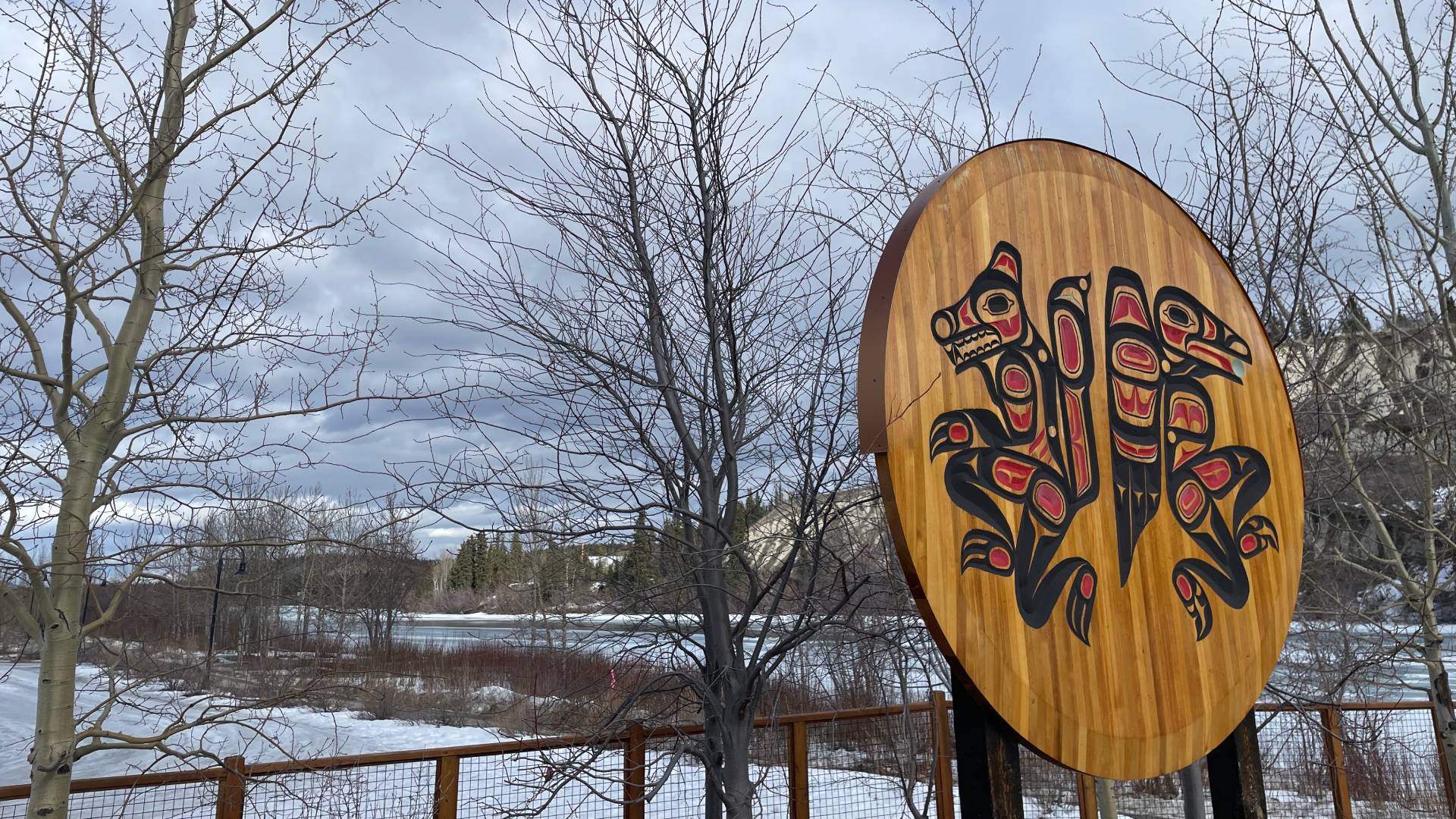 A wooden sign is depicted with a unique circular native design on it. The design features a bird prominently displayed. The sign is large, measuring 592 pixels wide and 850 pixels tall, positioned at coordinates (1132, 180) on a background that appears to be outdoors. The colors of the sign include shades of brown, gray, and blue. The surrounding environment includes a tree and branches, suggesting a park or natural setting. The overall scene conveys a sense of artistry and cultural significance, with a touch of whimsy from the bird motif.