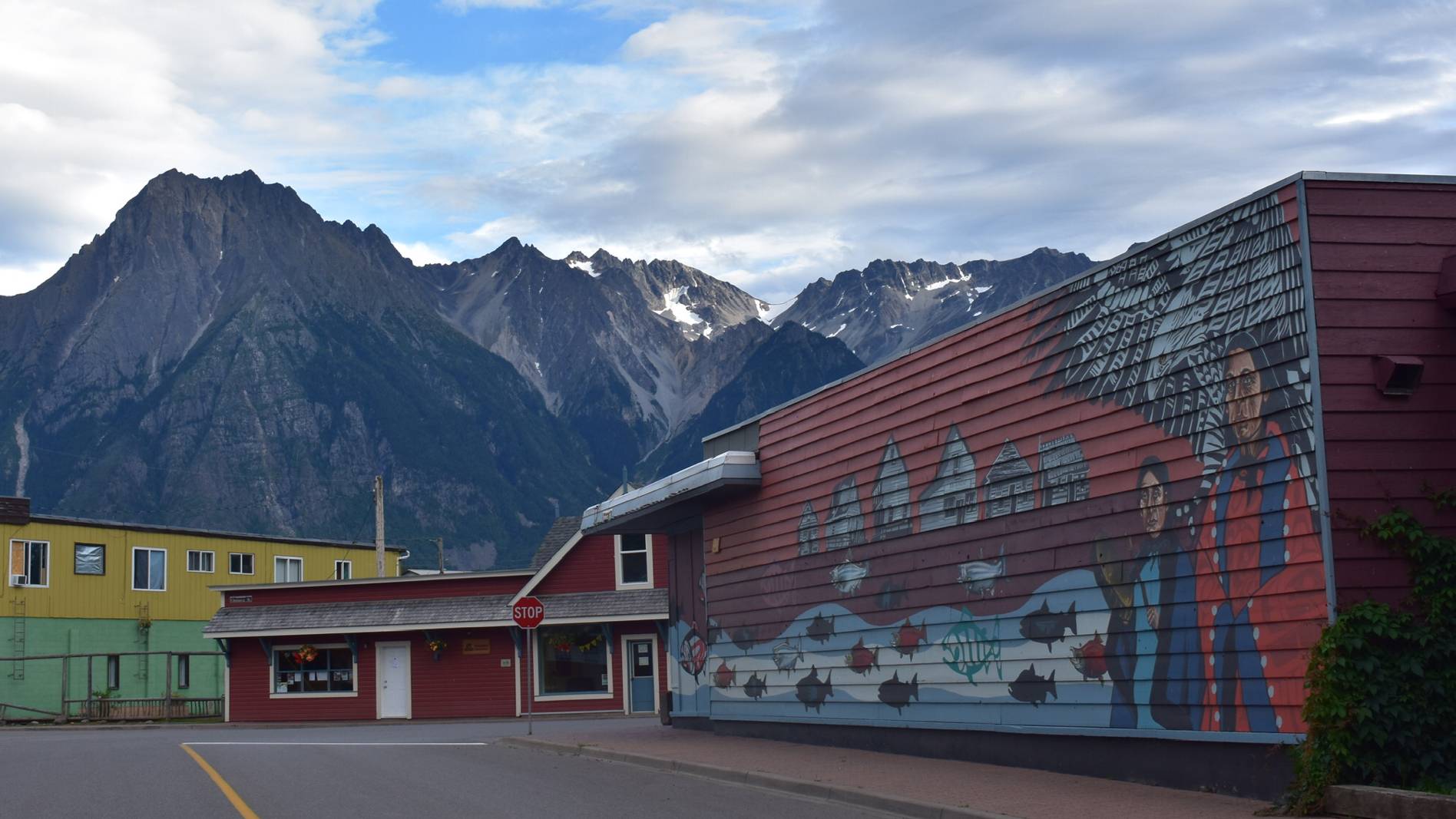 A picturesque street scene captured in this image showcases a vibrant building with a colorful facade standing out against a backdrop of majestic mountains. The architecture is diverse, with a stop sign visible in the foreground and a lamp post adding to the urban charm. The mountains in the distance provide a stunning natural contrast to the man-made structures in the foreground. The color palette is rich and varied, with shades of green, red, and blue dominating the scene. Overall, the image captures a harmonious blend of natural beauty and urban development.