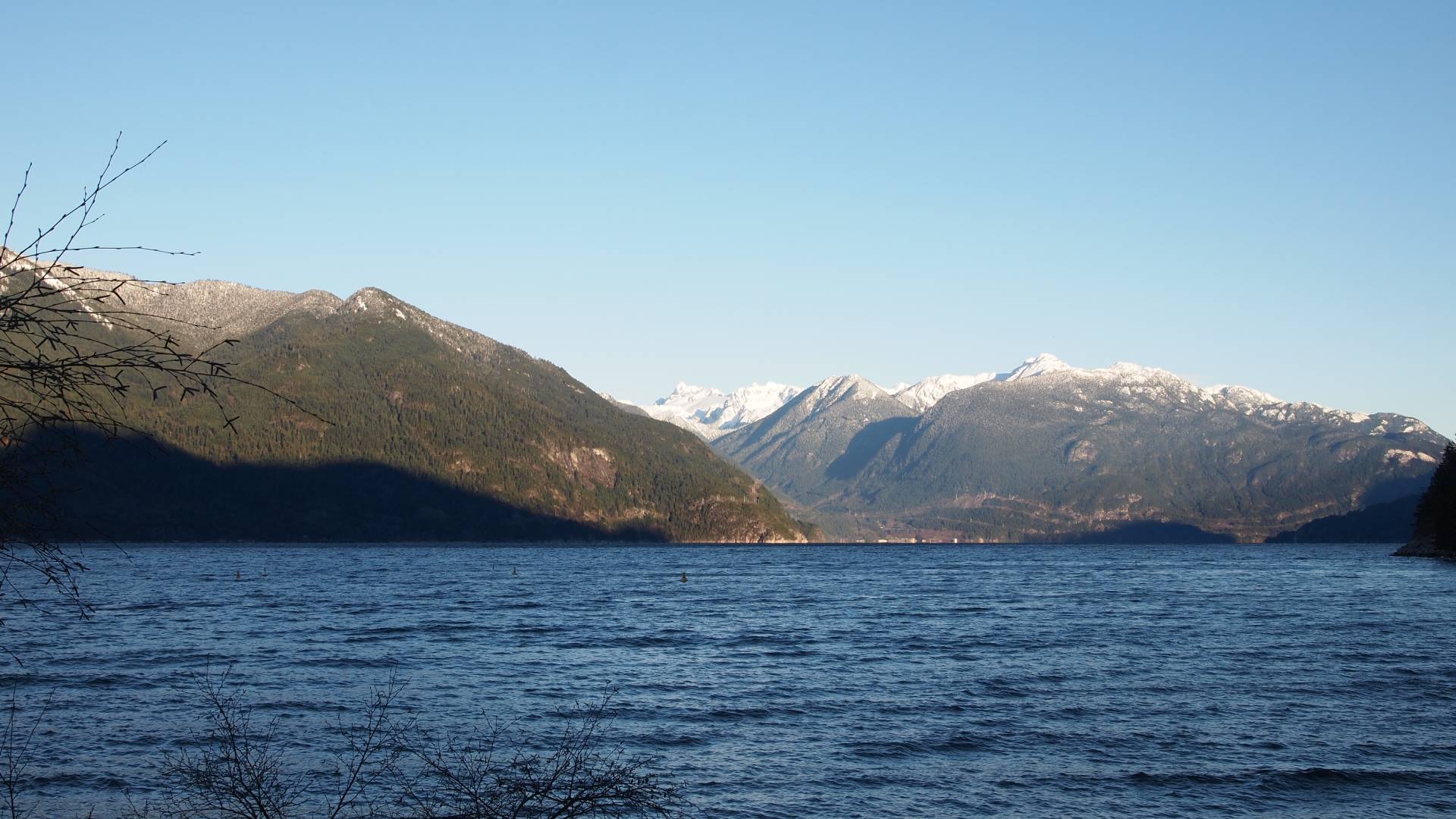 A serene scene of a lake nestled in between majestic mountains. The crystal-clear water reflects the towering peaks in the background, creating a breathtaking view. The mountains are partially covered in snow, adding a touch of elegance to the landscape. A few boats can be seen peacefully floating on the calm water, with a lone person swimming nearby. The blue hues of the lake contrast beautifully with the earthy tones of the mountains, creating a harmonious balance of colors. This tranquil setting showcases the beauty of nature and invites viewers to immerse themselves in the peaceful atmosphere.
